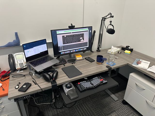 My office setup, with a MacBook Pro next to a large monitor with a Shure MV7x mic on a boom stand.